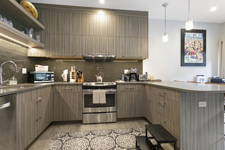 A modern L-shaped kitchen with wooden cabinets, stainless steel appliances, grey tiled backsplash, a patterned rug, and two pendant lights. A poster and small black stool are also visible.