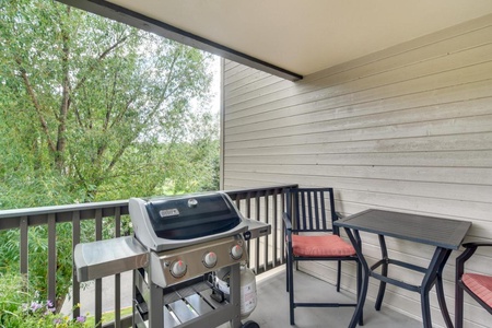 A covered balcony with a barbecue grill, and outdoor seating that includes a small table and two chairs. A leafy green view is visible through the railing.