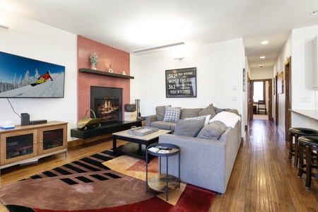 A modern living room with a gray sectional sofa, wooden floor, fireplace, wall-mounted TV displaying skiing, and hallway leading to other rooms. An area rug and coffee table are in the center.