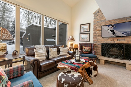 A cozy living room with a fireplace, leather sofa, armchairs, and a TV displaying a skier. Large windows reveal a snowy outdoor scene. The room features rustic and cabin-style decor.