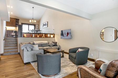 A modern living room features a gray sectional sofa, two gray armchairs, a leather armchair, and a wooden coffee table. A TV on the wall shows a skier. Open kitchen area with stairs in the background.