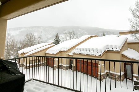 A snowy scene of a residential area with snow-covered roofs and driveways, viewed from a balcony with a black grill visible in the foreground. Background shows cloudy, snow-covered mountains.