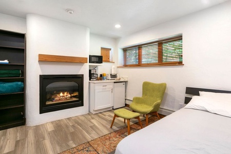 A cozy room with a fireplace, a green chair, a bed, and a small kitchenette with a microwave, coffee maker, and mini-fridge. The room has wooden floors and a window with blinds.