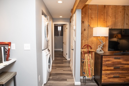 A hallway with a stackable washer and dryer, wooden walls, a TV on a dresser, and a sled leaning against it. A door and a small hallway visible at the end. Modern, rustic decor and lighting.