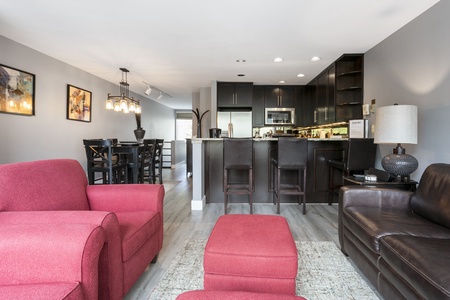 A modern, open-concept living area with red armchairs, a brown sofa, a dining table, and a kitchen with dark cabinets and bar stools.