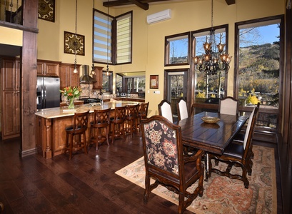 A spacious kitchen and dining area with wooden floors, a large central island with bar stools, decorative pendant lights, a long dining table with chairs, and large windows providing scenic views.