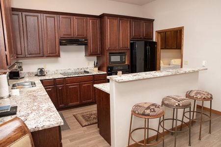 A kitchen with dark wooden cabinets, granite countertops, a black refrigerator, oven and microwave, and a breakfast bar with three cushioned stools.