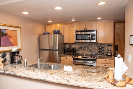 A kitchen with wooden cabinets, stainless steel appliances, a marble countertop, and various small appliances including a coffee maker and blender. A painting is hung on the wall.