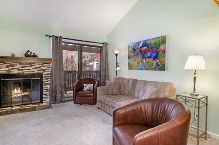 A cozy living room features a fireplace with a stone mantel, a beige sofa, two brown chairs, and a painting of a moose on the wall. A floor lamp and a table lamp provide additional lighting.