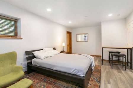 A bedroom with a wooden bed frame, white bedding, a green armchair, a tall mirror, a small desk with two stools, and recessed lighting. The room has wooden flooring and a colorful area rug.