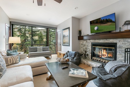 A cozy living room with a fireplace, a TV above it, a large window with a view of trees, two sofas, a bean bag chair, and a coffee table with magazines and a remote control.