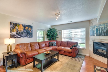 A living room with a large brown leather sectional sofa, coffee table, indoor plant, fireplace, and artwork on walls. Natural light enters through windows with blinds.