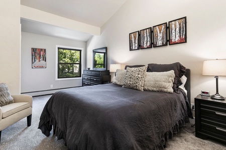 A bedroom with a large bed, dark bedding, two nightstands with lamps, a dresser with a mirror, wall art, and a window with a view of greenery.