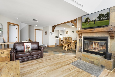 Living room with leather sofa, stone fireplace, TV showing golf, and wooden dining furniture leading to a kitchen. Natural light from windows highlights the wooden flooring and furnishings.