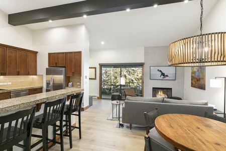 A modern open-plan kitchen and living area with wooden cabinets, granite countertops, a dining table, a couch facing a fireplace, and a large window offering a view of trees outside. A large ceiling light hangs above the room.