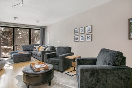 A living room featuring dark gray upholstered furniture, a light wood floor, framed pictures on the wall, and large windows letting in natural light.