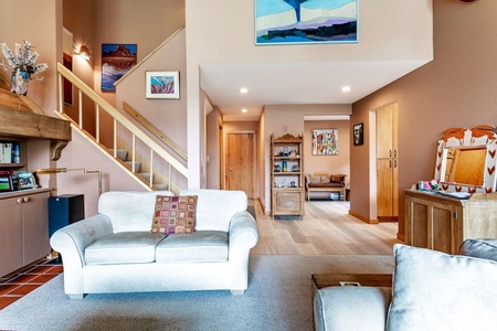 A cozy living room features neutral-colored furniture, framed artwork on the walls, a staircase leading to an upper level, and a mix of wooden and carpeted flooring.