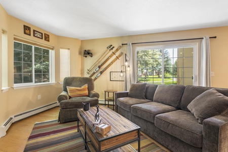 A living room with tan walls, a large dark green sofa, an armchair, a wooden coffee table, wall decor featuring skis, and two windows offering views of a grassy yard.