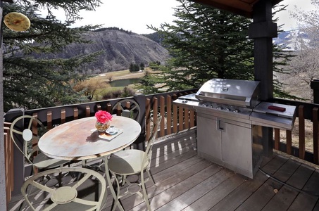 A wooden deck with a gas grill and a round metal table with three chairs and a vase of flowers, overlooking a hilly landscape with trees.