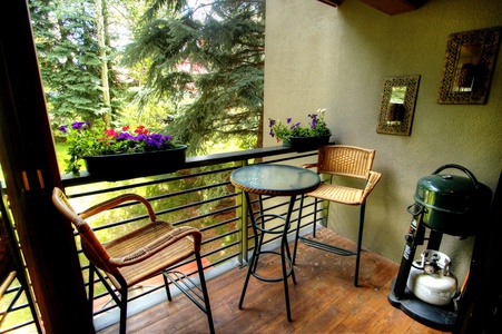 A small balcony with two wicker chairs, a round glass table, a barbecue grill, and flower boxes on the railing, surrounded by greenery.