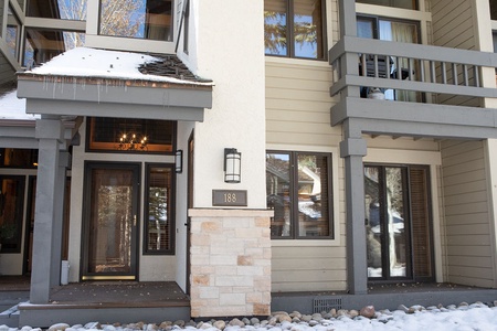 Exterior view of a two-story building with snow on the ground, showcasing wooden architectural details, a chandelier visible through the front glass door, and the number "188" displayed beside the entrance.