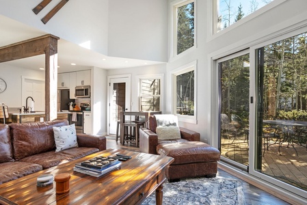 A modern living room with leather furniture, a wooden coffee table, large windows, and sliding glass doors opening to a patio. The kitchen is visible on the left with white cabinetry and stainless steel appliances.