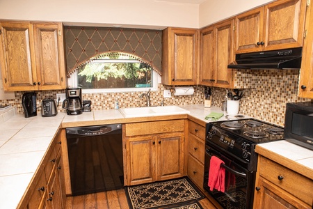 A cozy kitchen featuring wooden cabinets, tile backsplash, white countertops, and appliances including a dishwasher, stove, microwave, and coffee maker. A window with a valance overlooks the sink.