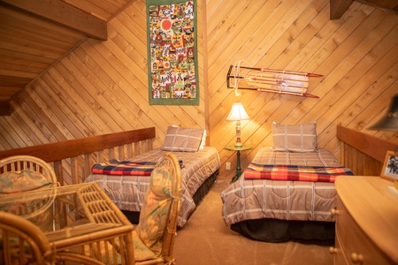 A cozy loft bedroom with two twin beds, a bedside table with a lamp, wooden walls, a wall hanging, vintage skis as decoration, and a wicker furniture set.