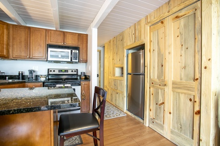 A kitchen with wooden cabinets, a black granite countertop, stainless steel appliances, and a wooden dining chair. A refrigerator is set into a wall with wooden sliding doors.