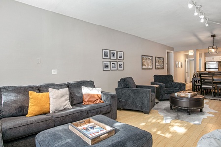 A living room with gray sofas, a round black coffee table, and wall art. A dining area and kitchen are visible in the background. The floor is hardwood with a cowhide rug near the seating area.