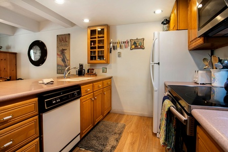 A kitchen with wooden cabinets, white appliances including a refrigerator, stove, and dishwasher, and a countertop with a sink and drying towel. Wall shelves hold glasses, and there is a rug on the floor.