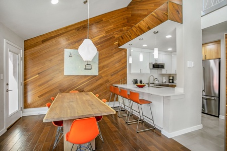 Modern kitchen and dining area with a wood-paneled accent wall, a wooden dining table, orange chairs, and white cabinetry. Stainless steel appliances and pendant lights are also present.