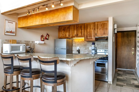 A modern kitchen with wooden cabinets, a granite countertop, a microwave, a refrigerator, an oven, and three bar stools. The space is well-lit with track lighting and has tiled flooring.