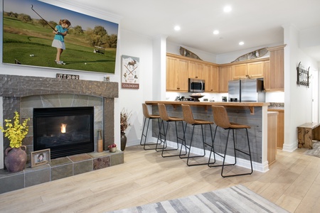 A modern kitchen with wooden cabinets, stainless steel appliances, and a central island with four barstools. There's a fireplace with a golf-themed painting above it and a rug on the wooden floor.