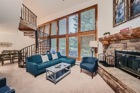 A spacious living room with high ceilings, large windows, a spiral staircase, blue sofas, and a stone fireplace. A dining area is visible in the background.