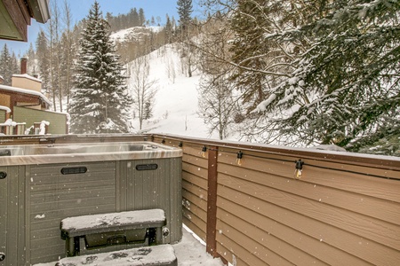 Snow-covered hot tub on a patio with a wooden fence, surrounded by snowy trees and hills in the background.