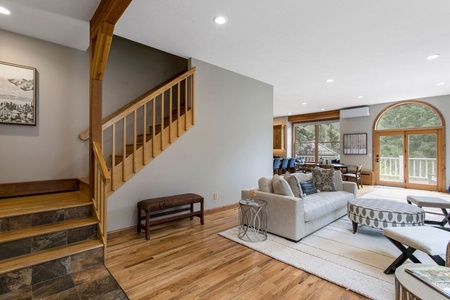 Living room with wooden floors, a staircase, gray sofa, round ottoman, and large windows leading to a balcony with a forest view.