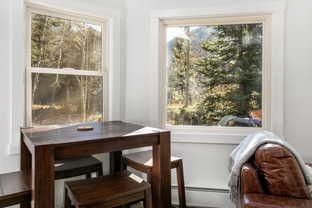 A small wooden dining table with three chairs is situated in front of two windows with scenic outdoor views. A leather chair with a blanket is partially visible on the right.