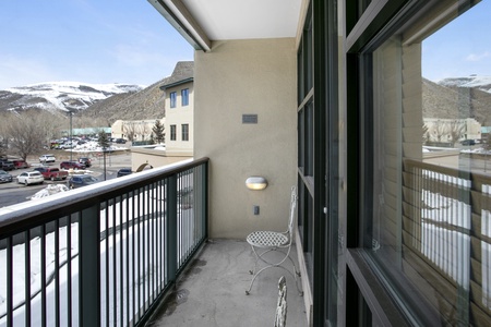 A small balcony with a metal chair overlooks a snowy street and parking area, with mountains visible in the background.