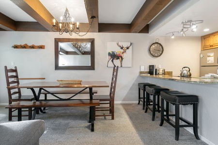 A dining area with a wooden table, bench, and chairs. A wall clock, mirror, and deer art are on the walls. A kitchen counter with three bar stools and appliances is partially visible in the background.