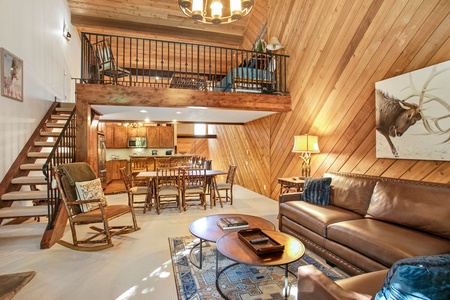 A living room with wood-paneled walls, a loft, leather couch, coffee tables, armchairs, and dining area. Stairs lead to the loft. Decor includes a cowboy picture and a bull painting.