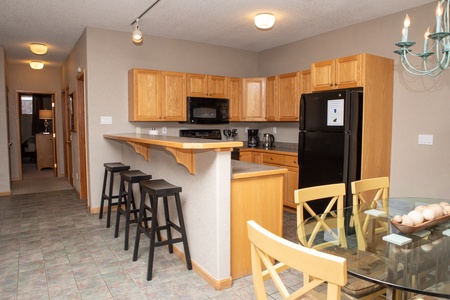 A modern kitchen with wooden cabinets, a black refrigerator, a stove, a microwave, a wooden breakfast bar with three black stools, a dining table with chairs, and a hallway leading to other rooms.