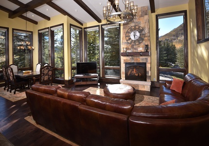 A living room features a brown leather sectional sofa, a stone fireplace with a clock above, large windows with scenic outdoor views, a chandelier, and a dining area with a wooden table and chairs.