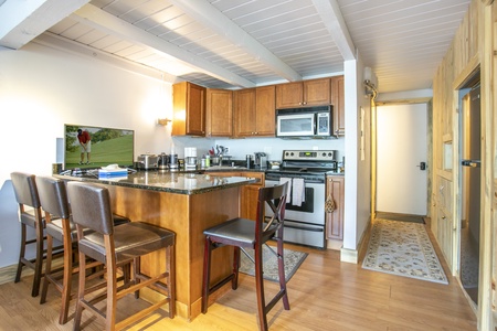 Kitchen with wooden cabinets, granite countertops, stainless steel appliances, and a breakfast bar with four chairs. A TV on the counter displays a scene of a person playing golf.