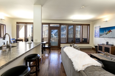 A modern living room with wooden floors features a gray sofa, a flat-screen TV displaying a snowboarder, a kitchen island with bar stools, and large windows with blinds providing a view of a balcony and outdoors.