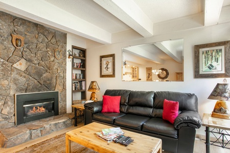 A cozy living room with a stone fireplace, leather couch with red pillows, wooden coffee table with magazines, bookshelves, and framed pictures on the wall.