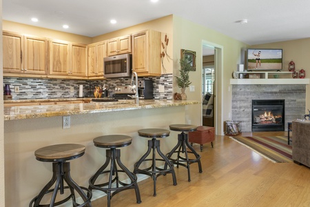 A kitchen with wooden cabinets, granite countertops, and four barstools. A living room with a fireplace and TV is visible in the background.