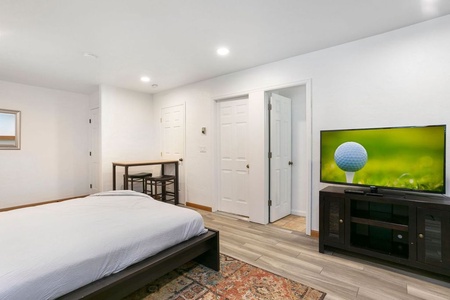 A bedroom with a white bed, a small table with two chairs, a TV displaying a golf ball on a stand, and a patterned rug on wood flooring.