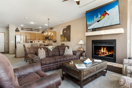 Cozy living room with brown sofas, a fireplace, wall-mounted TV showing a skier, and a coffee table. The kitchen with wooden cabinets is visible in the background. Decorative items adorn the space.