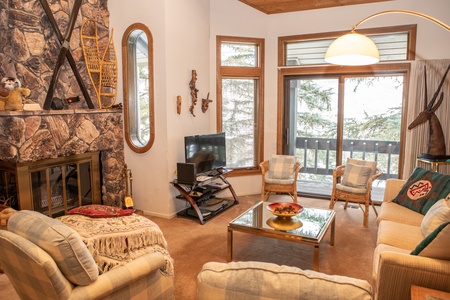 A cozy living room with a stone fireplace, TV, and glass coffee table. Multiple chairs and a sofa provide seating. Large windows and a sliding glass door offer views of a wooded area outside.